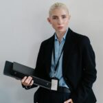 Positive independent businesswoman in black formal clothes standing with hand in pocket and folders in hand against white wall while looking at camera with confidence